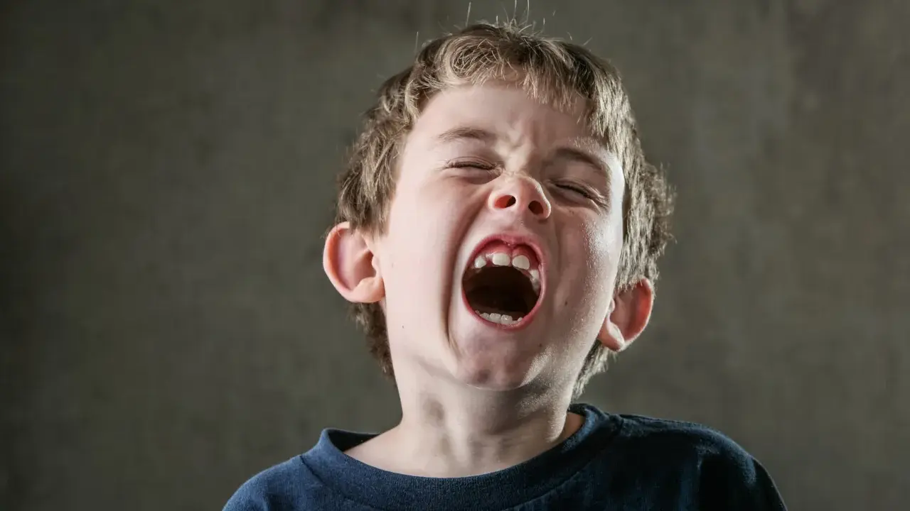 Ein Junge schreit mit weit geöffnetem Mund und geschlossenen Augen. Sein Gesichtsausdruck zeigt ein starkes, impulsives Verhalten, das durch die Intensität seines Rufs unterstrichen wird.