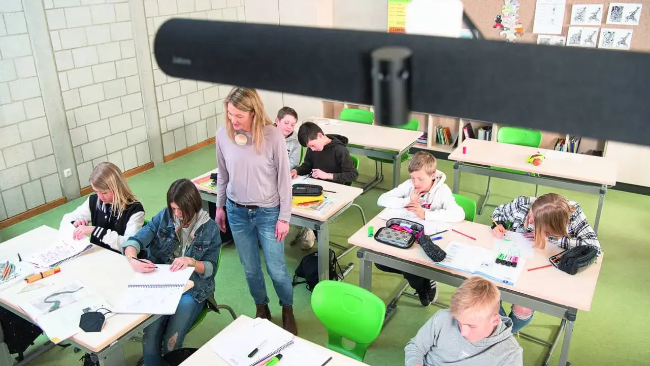 Lehrerin betreut Schüler im Klassenzimmer, die an ihren Tischen arbeiten. Ein modernes Setup für **hybrid unterricht**.