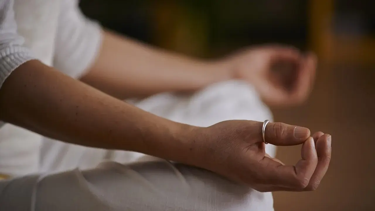 Eine Person in weißer Kleidung meditiert, die Hände im Schoß gefaltet. Ein silberner Ring ziert den Finger. Es herrscht eine tiefe ruhe im klassenzimmer.