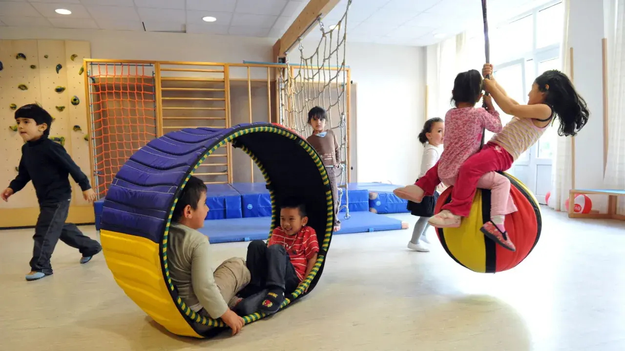 Kinder mit großem Bewegungsdrang toben in einer Turnhalle. Ein Junge rollt in einer Röhre, während andere auf einem großen Ball schaukeln.