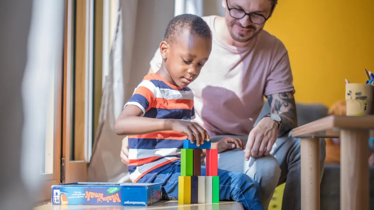 Ein Mann spielt mit einem Kind im Kindergarten. Der Junge baut einen Turm aus bunten Holzklötzen. Der Mann lächelt ihn an. Kein Trennungsschmerz im Kindergarten, nur Spiel und Spaß.