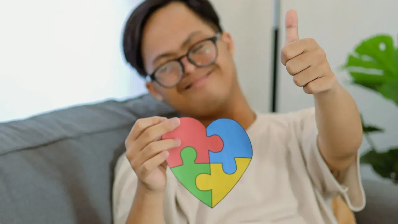 Ein Junge mit Down-Syndrom hält ein Herz aus Puzzleteilen, das für Autismus-Tests bei Kindern ab 7 Jahren stehen könnte. Er gibt einen Daumen hoch.
