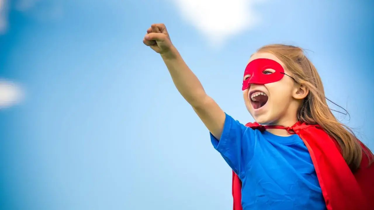 Ein kleines Mädchen im Superheldenkostüm mit rotem Cape und Maske jubelt unter blauem Himmel. Ein guter Start in der neuen Schule!