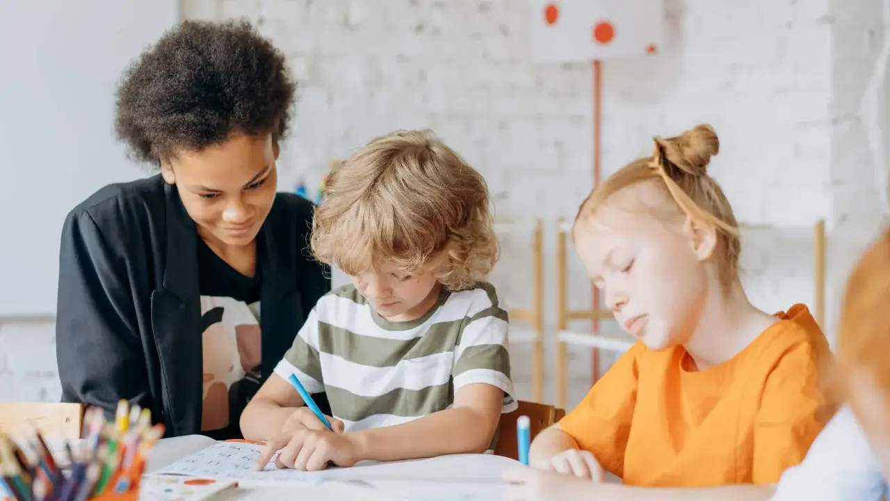 Kinder konzentriert bei Lernritualen in der Grundschule. Ein Mädchen mit schwarzer Afro-Frisur schaut zu, während ein Junge mit blonden Locken und ein Mädchen mit roten Haaren schreiben.