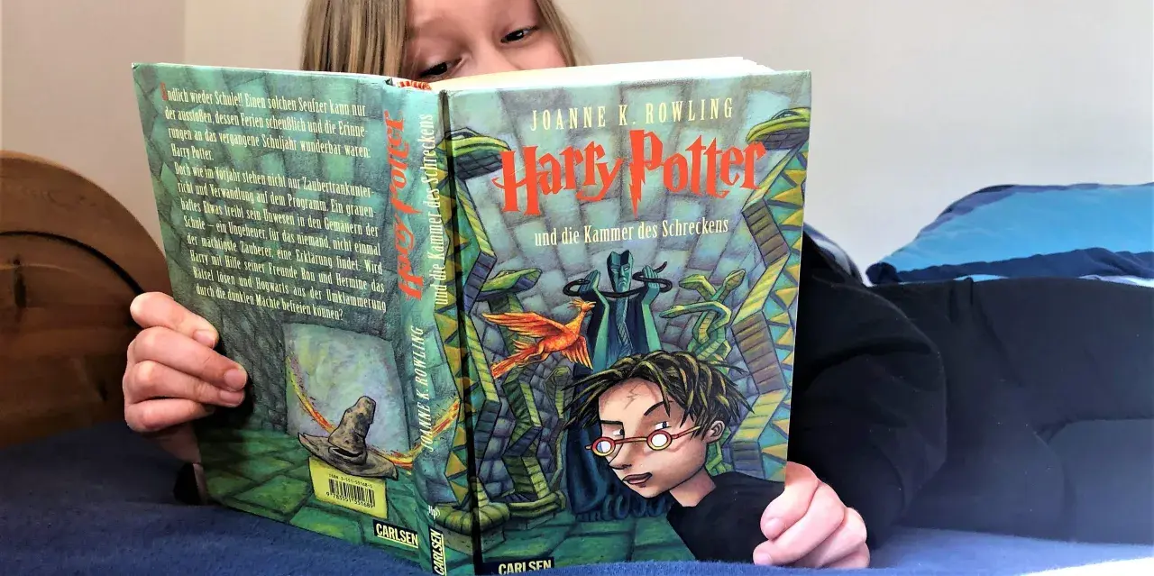 Ein Kind liest Harry Potter und die Kammer des Schreckens, eine Harry Potter Kinderversion. Der Zauberhut und ein Phönix sind auf dem Cover zu sehen.