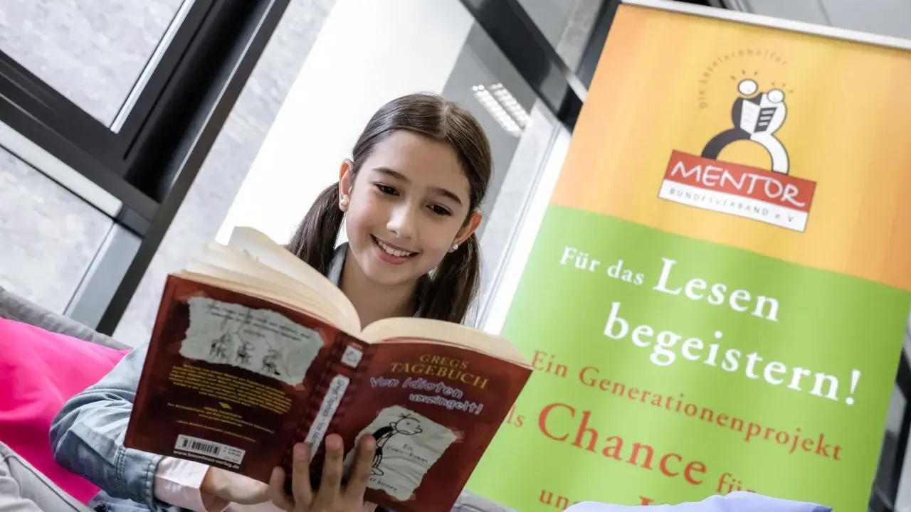 Ein lächelndes Mädchen liest ein Buch. Ein Banner im Hintergrund wirbt für "MENTOR" und "digitales lernen" durch Lesen.