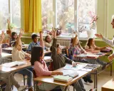 Grundschüler melden sich eifrig, ihre Sitzordnung zur Wand hin ausgerichtet, während die Lehrerin lächelt.