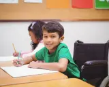 Ein Junge im Rollstuhl lächelt, während er im Unterricht schreibt. Dies ist ein Beispiel für **Inklusion in der Schule**.