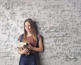 Ein lächelndes Mädchen mit Büchern vor einer Wand voller mathematischer Formeln. Sie zeigt, wie kann man sich in Mathe verbessern: durch Lernen und Üben.