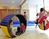 Kinder mit großem Bewegungsdrang toben in einer Turnhalle. Ein Junge rollt in einer Röhre, während andere auf einem großen Ball schaukeln.
