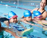 Fröhliche Kinder mit blauen Badekappen üben das schwimmen in der Grundschule. Eine Lehrerin hilft ihnen im Wasser.
