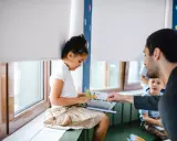 Ein Mädchen sitzt am Fenster und liest ein Buch. Ein Mann zeigt auf eine Seite. Dies fördert eigenverantwortliches Lernen.