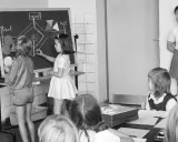 Unterricht in einer Schule um 1900. Kinder lernen an einer Tafel, während die Lehrerin zusieht.