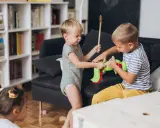 Zwei kleine Jungen streiten sich um ein grünes Stofftier. Einer hält einen Kochlöffel, der andere zieht am Spielzeug. Ein kleines Kind schaut zu.