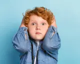 Rotes Lockenköpfchen mit weit aufgerissenen Augen hält sich die Ohren zu, wirkt überfordert. Ein Ausdruck von motorischer Unruhe bei Kindern.