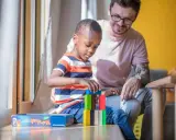 Ein Mann spielt mit einem Kind im Kindergarten. Der Junge baut einen Turm aus bunten Holzklötzen. Der Mann lächelt ihn an. Kein Trennungsschmerz im Kindergarten, nur Spiel und Spaß.