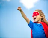 Ein kleines Mädchen im Superheldenkostüm mit rotem Cape und Maske jubelt unter blauem Himmel. Ein guter Start in der neuen Schule!
