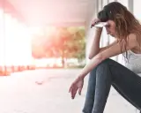 Junge Frau sitzt nachdenklich da, Handy auf der Stirn. Perfektionismus und Versagensangst belasten sie.