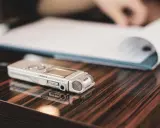 Digitaler Rekorder liegt auf glänzendem Holz, bereit für online diktate. Ein aufgeschlagenes Notizbuch im Hintergrund.
