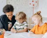 Kinder konzentriert bei Lernritualen in der Grundschule. Ein Mädchen mit schwarzer Afro-Frisur schaut zu, während ein Junge mit blonden Locken und ein Mädchen mit roten Haaren schreiben.
