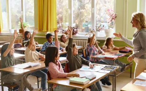Grundschüler melden sich eifrig, ihre Sitzordnung zur Wand hin ausgerichtet, während die Lehrerin lächelt.