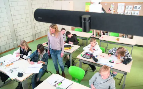 Lehrerin betreut Schüler im Klassenzimmer, die an ihren Tischen arbeiten. Ein modernes Setup für **hybrid unterricht**.