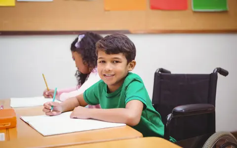 Ein Junge im Rollstuhl lächelt, während er im Unterricht schreibt. Dies ist ein Beispiel für **Inklusion in der Schule**.