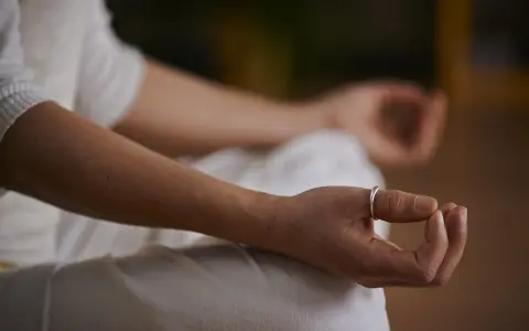 Eine Person in weißer Kleidung meditiert, die Hände im Schoß gefaltet. Ein silberner Ring ziert den Finger. Es herrscht eine tiefe ruhe im klassenzimmer.