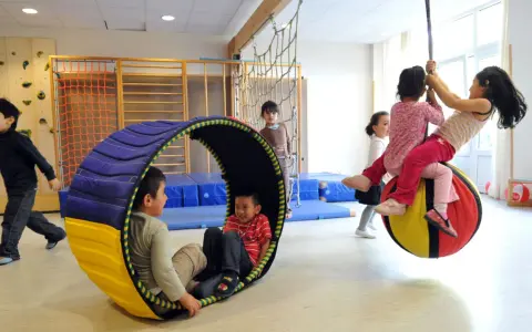 Kinder mit großem Bewegungsdrang toben in einer Turnhalle. Ein Junge rollt in einer Röhre, während andere auf einem großen Ball schaukeln.