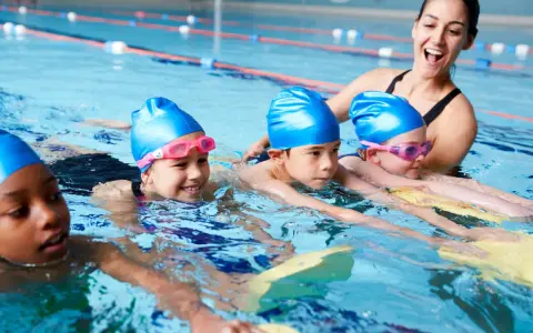 Fröhliche Kinder mit blauen Badekappen üben das schwimmen in der Grundschule. Eine Lehrerin hilft ihnen im Wasser.