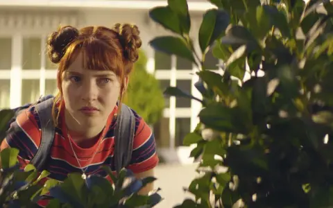 Ida aus "Die Schule der magischen Tiere" späht neugierig hinter Büschen hervor.