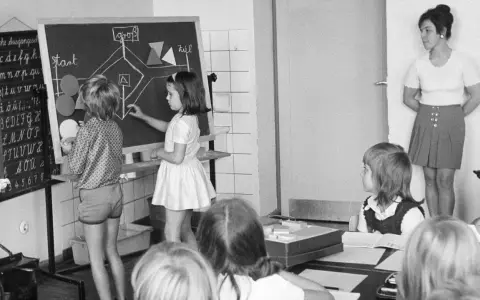 Unterricht in einer Schule um 1900. Kinder lernen an einer Tafel, während die Lehrerin zusieht.