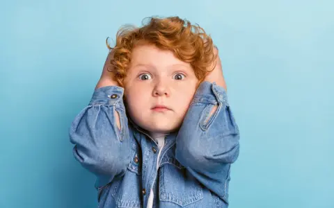 Rotes Lockenköpfchen mit weit aufgerissenen Augen hält sich die Ohren zu, wirkt überfordert. Ein Ausdruck von motorischer Unruhe bei Kindern.