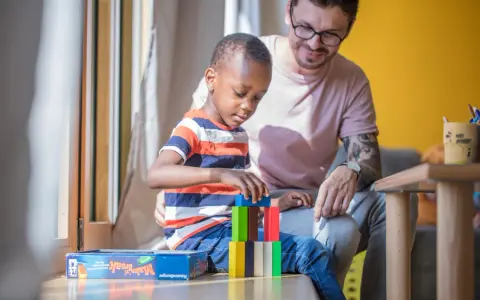 Ein Mann spielt mit einem Kind im Kindergarten. Der Junge baut einen Turm aus bunten Holzklötzen. Der Mann lächelt ihn an. Kein Trennungsschmerz im Kindergarten, nur Spiel und Spaß.