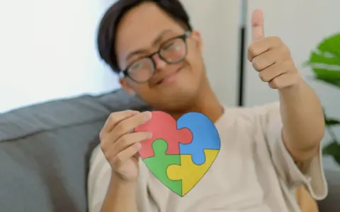Ein Junge mit Down-Syndrom hält ein Herz aus Puzzleteilen, das für Autismus-Tests bei Kindern ab 7 Jahren stehen könnte. Er gibt einen Daumen hoch.