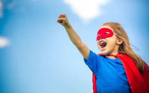 Ein kleines Mädchen im Superheldenkostüm mit rotem Cape und Maske jubelt unter blauem Himmel. Ein guter Start in der neuen Schule!