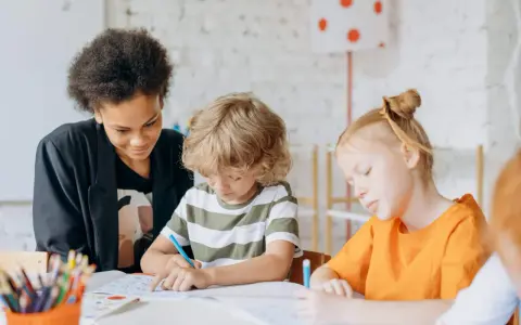 Kinder konzentriert bei Lernritualen in der Grundschule. Ein Mädchen mit schwarzer Afro-Frisur schaut zu, während ein Junge mit blonden Locken und ein Mädchen mit roten Haaren schreiben.