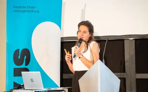 Eine Rednerin spricht über den Umgang mit Misserfolg. Im Hintergrund ein Banner der Deutschen Kinder- und Jugendstiftung.