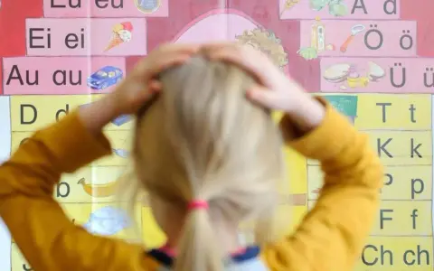Ein Kind mit Händen am Kopf vor einer Buchstabenwand, die auf Schwierigkeiten beim Lesen und Schreiben (LRS) bei Kindern hindeuten könnte.