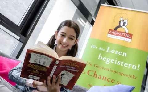 Ein lächelndes Mädchen liest ein Buch. Ein Banner im Hintergrund wirbt für "MENTOR" und "digitales lernen" durch Lesen.