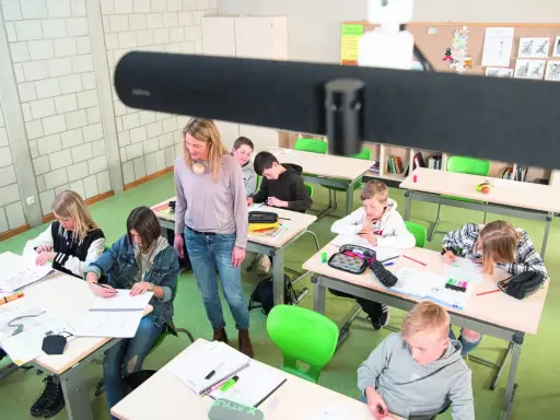 Lehrerin betreut Schüler im Klassenzimmer, die an ihren Tischen arbeiten. Ein modernes Setup für **hybrid unterricht**.