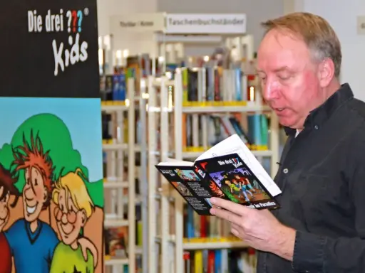Ulf Blank liest ein Buch der "Die drei ??? Kids" in einer Bibliothek.
