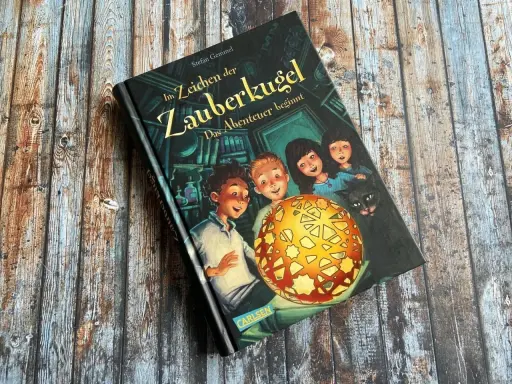 Ein Buchcover mit dem Titel "Im Zeichen der Zauberkugel". Kinder und eine Katze blicken neugierig auf eine leuchtende Kugel.