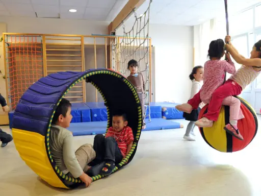 Kinder mit großem Bewegungsdrang toben in einer Turnhalle. Ein Junge rollt in einer Röhre, während andere auf einem großen Ball schaukeln.