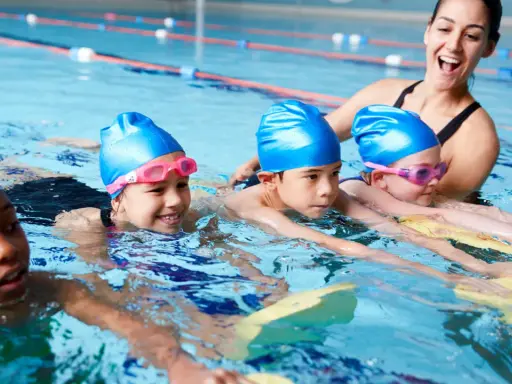 Fröhliche Kinder mit blauen Badekappen üben das schwimmen in der Grundschule. Eine Lehrerin hilft ihnen im Wasser.