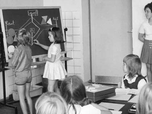 Unterricht in einer Schule um 1900. Kinder lernen an einer Tafel, während die Lehrerin zusieht.