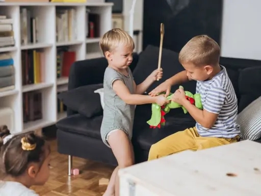 Zwei kleine Jungen streiten sich um ein grünes Stofftier. Einer hält einen Kochlöffel, der andere zieht am Spielzeug. Ein kleines Kind schaut zu.