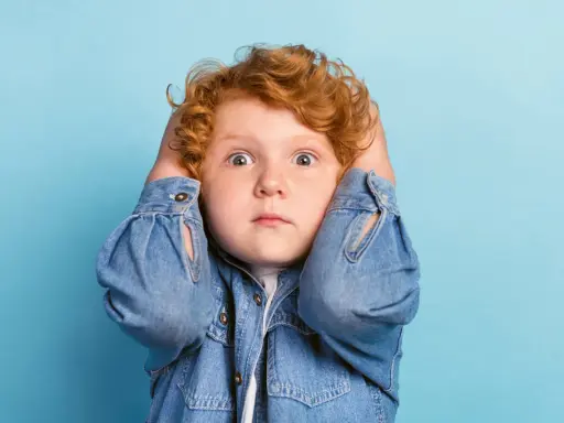 Rotes Lockenköpfchen mit weit aufgerissenen Augen hält sich die Ohren zu, wirkt überfordert. Ein Ausdruck von motorischer Unruhe bei Kindern.