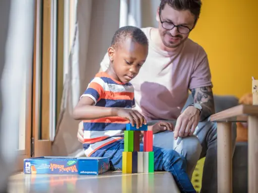 Ein Mann spielt mit einem Kind im Kindergarten. Der Junge baut einen Turm aus bunten Holzklötzen. Der Mann lächelt ihn an. Kein Trennungsschmerz im Kindergarten, nur Spiel und Spaß.