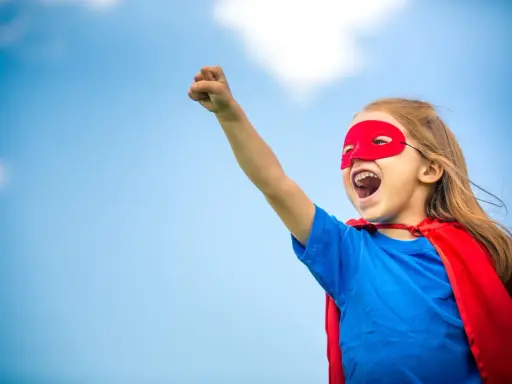 Ein kleines Mädchen im Superheldenkostüm mit rotem Cape und Maske jubelt unter blauem Himmel. Ein guter Start in der neuen Schule!